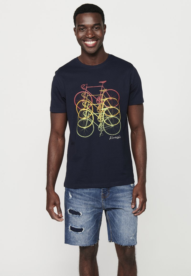 Camiseta de algodón azul marino con un diseño gráfico colorido de una bicicleta, de mangas cortas y cuello redondo. Combinada con unos pantalones cortos de denim desgastados.