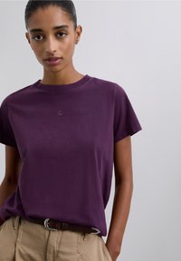 Paars katoenen T-shirt met een ronde hals en korte mouwen. Kleine logodetail op de borst, gecombineerd met beige broek en een bruine riem.