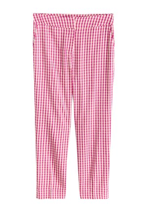 Jonge vrouw met lang haar, dragend een roze-wit geruit mouwloos topje en bijpassende broek, staand tegen een roze muur met beige platte schoenen.