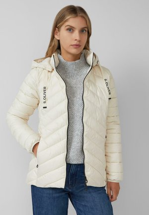 Lichte crème pufferjack met capuchon, gewatteerd ontwerp, zwarte ritsluiting en S. Oliver-logo op de mouwen, gedragen over een grijze sweater.