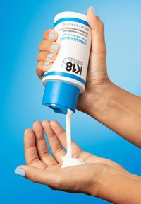 Witte crème komt uit een cilindervormige fles met een blauwe dop, gelabeld "K8 Damage Shield," die boven een open hand tegen een blauwe achtergrond wordt gehouden.