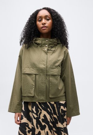 Femme aux cheveux bouclés portant une veste à capuche vert olive et une jupe beige avec des motifs abstraits noirs, debout devant un fond uni.