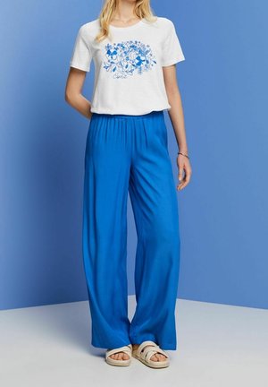 Camiseta blanca recortada con estampado floral azul, combinada con pantalones sueltos azules y sandalias ligeras. Colores sólidos y corte relajado.