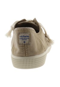 Zapatillas de lona beige con una superficie texturizada, puntera redonda y suela de goma. Presenta una etiqueta azul en la parte posterior y acentos de borlas en los cordones.