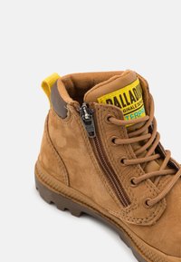 Bottine en suède marron avec une étiquette de logo jaune, une fermeture éclair sur le côté, des lacets marron et une semelle en caoutchouc texturée. Comprend un col rembourré et des surpiqûres décoratives.