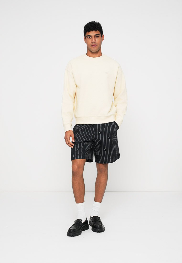 Homme debout portant un sweat-shirt crème, un short noir à rayures avec de petits motifs blancs, des chaussettes blanches et des mocassins noirs.