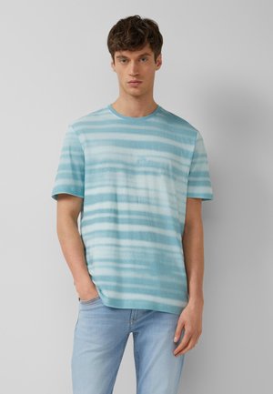 Jeune homme portant un t-shirt à manches courtes rayé bleu et blanc et un jean bleu clair, debout avec une main dans la poche.