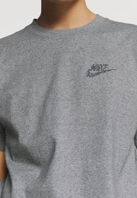 Šedé tričko s krátkým rukávem vyrobené z bavlny, s barevným, třpytivým logem Nike na levé části hrudi. Hladká textura s klasickým střihem.