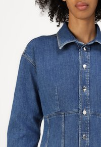 Denimkjole med krage, sølvknapper og strukturerte sømmer. Stoffs sammensetning har en lett falmet blåfarge og en glatt tekstur.