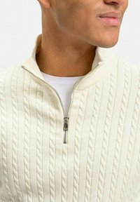 Krämfärgad kabelstickad tröja med hög krage och silverfärgad dragkedja. Har vertikal ribbing och texturerade mönster genom hela plagget.