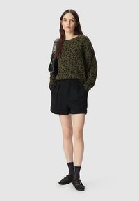 Olive green leopard print knit sweater, black pleated shorts, black socks, and shoes. Model standing with hands in pockets.