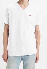 T-shirt V-cou à manches courtes en coton blanc avec un petit patch logo rouge Levi's sur la poitrine gauche. Texture douce, coupe décontractée.