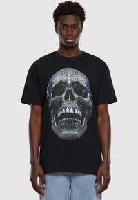 Černé bavlněné tričko s výraznou grafikou lebky s metalickými detaily, velkými očními jamkami a ostrými zuby. Pohodlný střih.