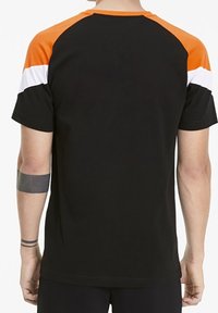 Hombre que lleva una camiseta negra de manga corta con hombros naranjas y una franja blanca a lo largo de las mangas, visto desde detrás.