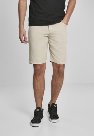 Beige bomuldsshorts med lommer, knælængde, båret med en sort t-shirt og sorte sneakers med hvide såler.