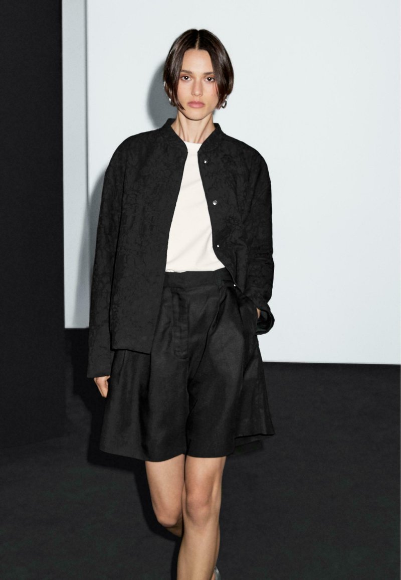 Zwarte textuur jacket met drukknoopjes, gedragen over een witte shirt en zwarte geplooide shorts. Soepele stof met een ontspannen pasvorm.