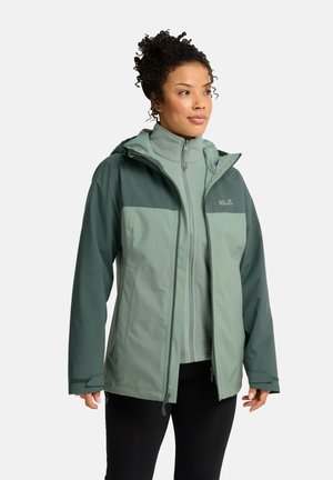 Vrouw met krullend haar die een lichtgroene en donkergroene outdoor jas met rits en zwarte broek draagt, staand tegen een effen achtergrond.
