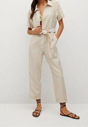 Beżowy kombinezon na guziki z krótkimi rękawami, wiązany w pasie i luźnym krojem. Posiada przednie kieszenie i jest zestawiony z czarnymi sandałami na paski.