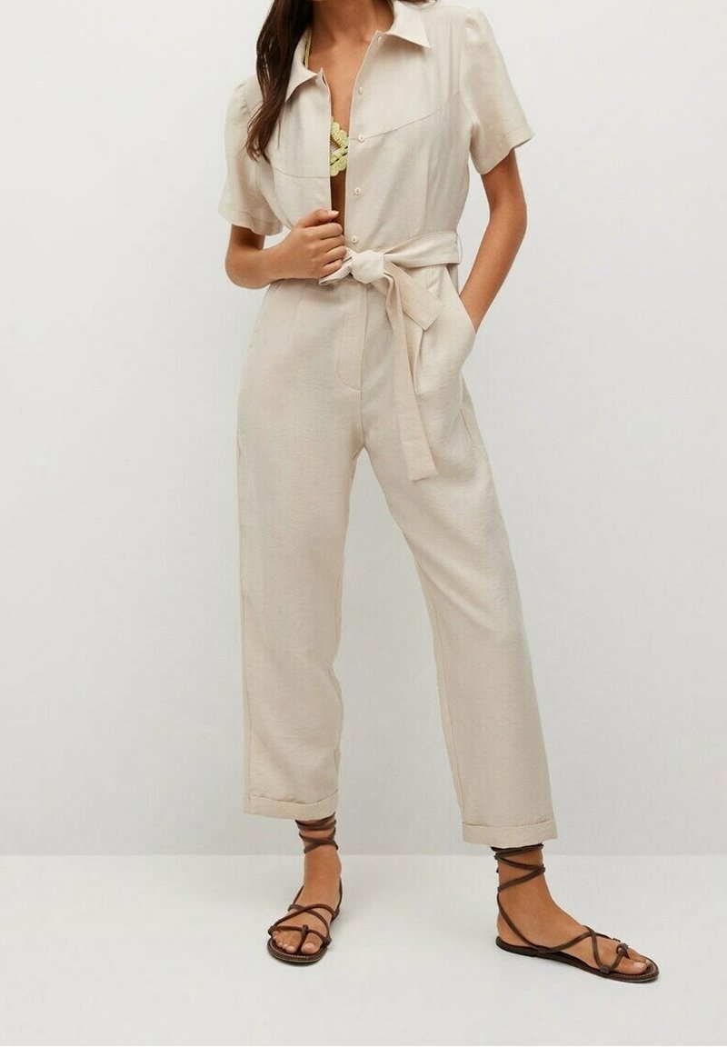 Combinaison beige à boutons sur le devant, manches courtes, taille ceinturée et coupe décontractée. Dotée de poches avant et portée avec des sandales noires à lanières.