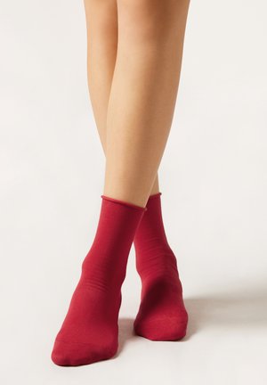 Piernas con calcetines rojos hasta el tobillo cruzadas sobre un fondo claro y liso, mostrando el ajuste y la textura de los calcetines.