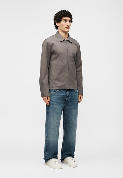 Άνδρας που φοράει γκρι μπουφάν με φερμουάρ, μπλε τζιν και λευκά αθλητικά, μπροστά από ένα απλό ανοιχτό φόντο.