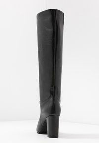 Bota de cuero negra hasta la rodilla con una textura lisa y suave, punta redondeada y tacón alto apilado. Costuras decorativas a lo largo de la parte posterior.