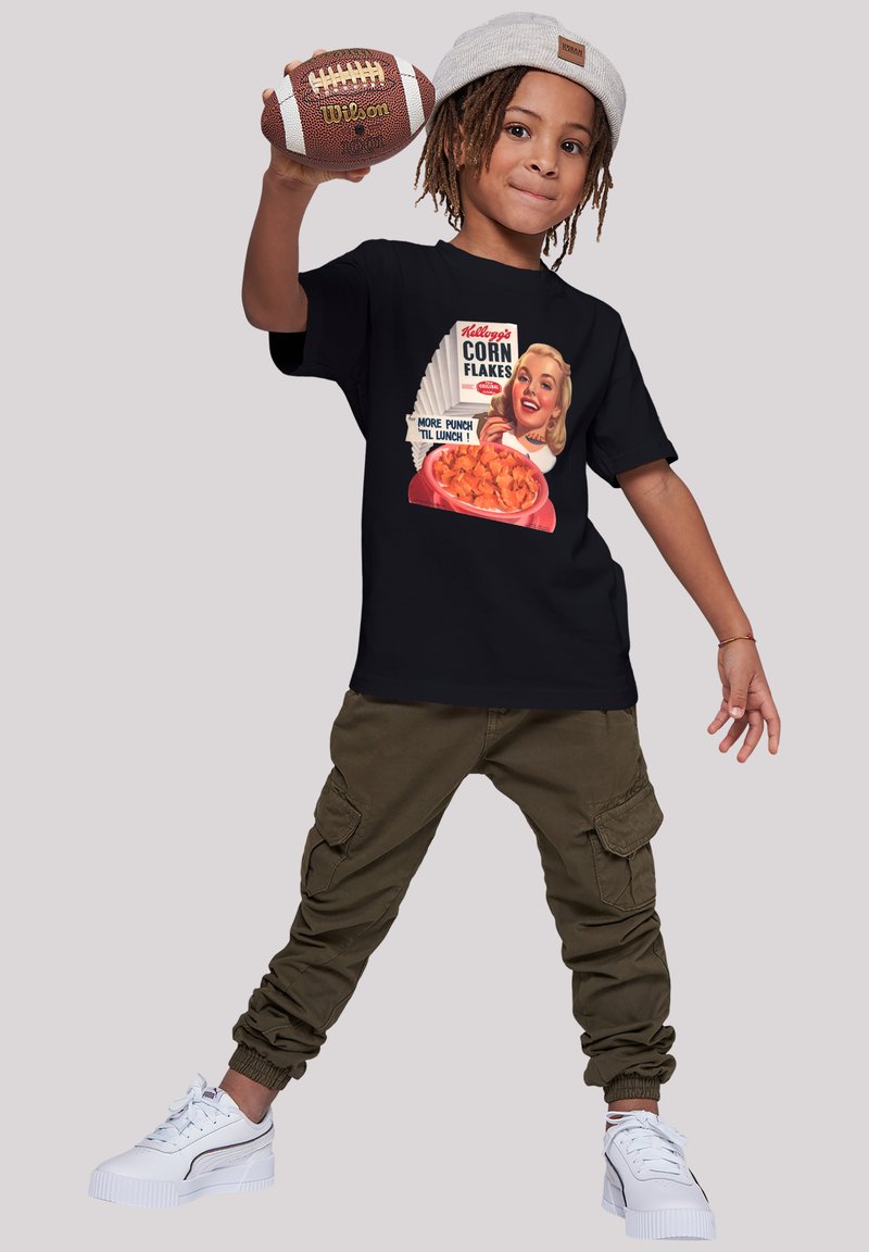Black t-shirt with retro cereal graphic, olive green cargo pants, and white sneakers. Child holding a brown football, wearing a light gray beanie.