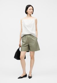 Γυναίκα που φοράει λευκό τοπ χωρίς μανίκια, χακί σορτς μήκους μέχρι το γόνατο, μαύρες μπαλαρίνες με μυτερή μύτη, κρατώντας μαύρη τσάντα, στέκεται μπροστά σε απλό φόντο.