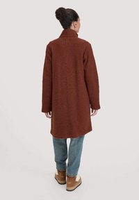 Cappotto in lana marrone, texturizzato, con colletto alto, maniche lunghe e spacchi laterali, abbinato a jeans azzurri chiari e stivali marroni.