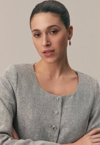 Tânără cu părul prins la spate, purtând o bluză gri cu model în chevron, cu nasturi decorativi și cercei de aur în formă de cerc.