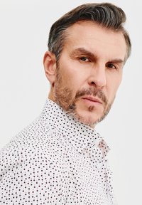 Biała koszula z małymi, ciemnoczerwonymi wzorami kwiatowymi, z kołnierzykiem zapinanym na guziki oraz teksturowanym materiałem, podkreślająca dopasowany krój i subtelne detale wzornicze.