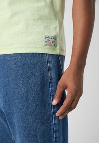 Tricou din bumbac de culoare verde deschis, cu mâneci scurte, având o etichetă la margine. Asortat cu blugi din denim albastri, evidențiind textura țesăturii.