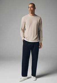 Camisa de manga larga texturizada color beige claro con cuello redondo, combinada con pantalones oscuros y zapatillas blancas. Diseño sencillo y moderno.
