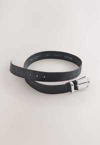 Czarny skórzany pasek z tłoczonym kwiatowym wzorem i srebrną klamrą, oznaczony „Next” i „Medium”, zwinięty na jasnym tle.