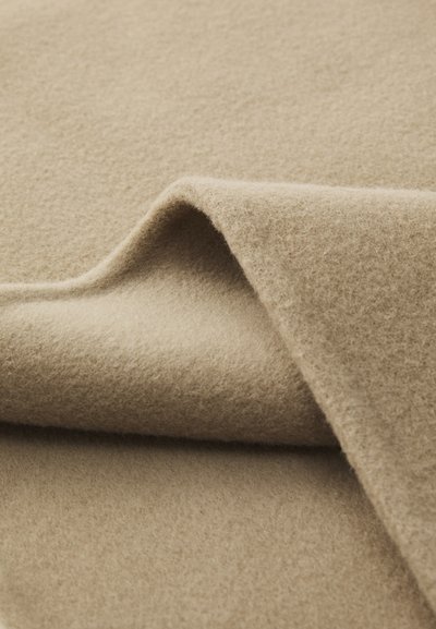 Tela de lana beige con una superficie suave y texturizada, parcialmente doblada para mostrar capas, destacando su color cálido y acabado liso.
