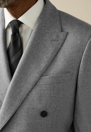 Gray double-breasted suit jacket with checked pattern, peak lapels, and a black button. Accompanied by a white shirt and a black tie with gold stripe.