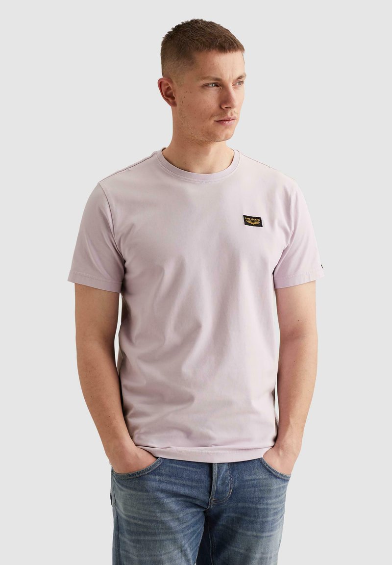 Jeune homme aux cheveux courts portant un t-shirt rose clair avec un petit écusson logo sur la poitrine et un jean bleu, mains dans les poches, regardant vers la droite.