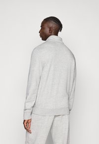 Ανοιχτό γκρι, μακρυμάνικο πουλόβερ με ψηλό γιακά και ριμπ μανσέτες. Ομαλή υφή, άνετη γραμμή, εμφανές ταιριαστό παντελόνι στη μέση.