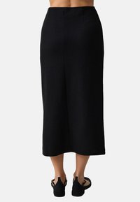 Jupe crayon noire de mi-longueur avec une texture lisse et des détails de couture subtils ; portée avec des chaussures noires à lanières et à semelle plate.