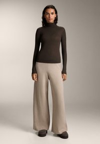 Dunkelbrauner gerippter Rollkragenpullover, beiger weit geschnittener Hose und grauen Plateauschuhen. Glatte Texturen mit einem enganliegenden Oberteil und lockerem Unterteil.