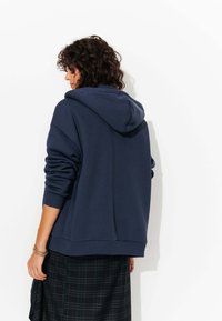 Sweat-shirt à capuche bleu marine avec des manches et un ourlet côtelés. Présente une coupe décontractée, une texture douce, et est associé à une jupe à carreaux sombre.