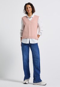 Kvinna som står, iklädd en ljusrosa ärmlös tröja över en vit skjorta med uppvikta ärmar, blåjeans och vita sneakers.