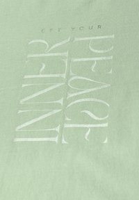 Vaaleanvihreä kangas, jossa on kohokuvioitu valkoinen teksti, joka lukee "SAAVUTA SISÄINEN RAUHASI." Silkkinen pinta, joka näyttää kevyeltä.