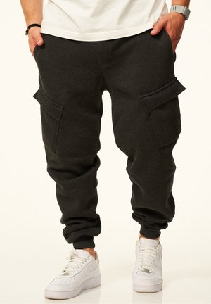 Person steht und trägt dunkelgraue Cargo-Jogger-Hosen und weiße Sneakers mit den Händen in den Taschen vor einem einfarbigen Hintergrund.