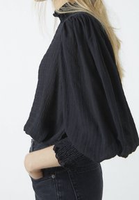 Schwarze, strukturierte Bluse mit Puffärmeln und gerafften Manschetten, mit einem hohen Kragen. Getragen mit dunklen Jeans, was den lässigen Stil betont.