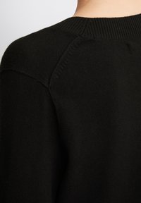 Schwarzer Strickpullover mit geripptem Ausschnitt, zeigt die Rückensaumnaht an der Schulter und eine glatte Textur. Minimalistisches Design ohne zusätzliche Muster.