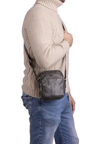 Borsa a tracolla in pelle nera texturizzata, con tasca con zip e tracolla regolabile. Design compatto e quadrato, caratterizzato da un logo sottile.
