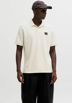 Jauns vīrietis, tērpies ziloņu kaula krāsas īsroku polo kreklā ar uzšuvumu uz krūtīm, melnās, platu kāju biksēs un tumšu beisbola cepuri, skatās uz sāniem.