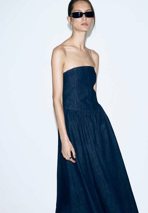 Kobieta ubrana w ciemną sukienkę z jeansu bez ramiączek i czarne okulary przeciwsłoneczne, stojąca na jednolitym jasnym tle.