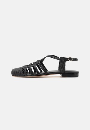 Sort flet-skolet læder flad sandal med flere remme, ankelspænde og lukket tå, vist fra siden på en hvid baggrund.
